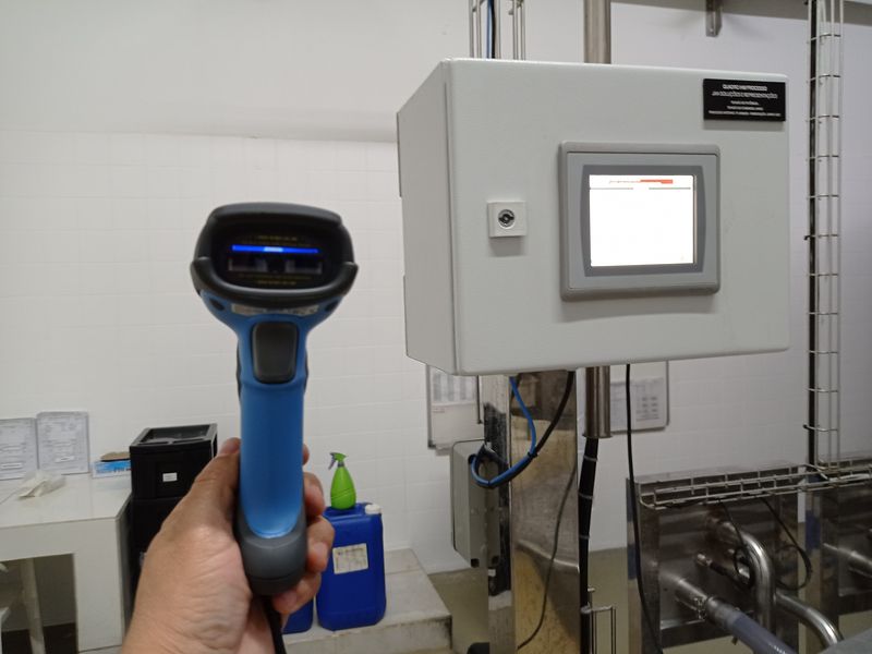 Leitor de código de barras na mão de um operador, em frente ao quadro de IHM do processo